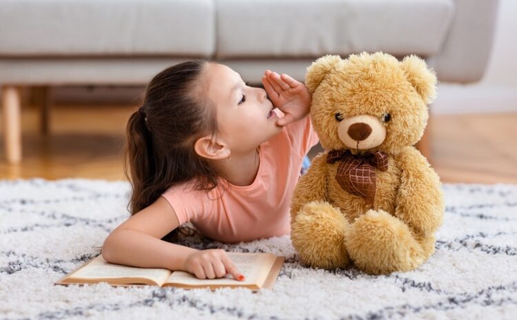 Kid’s Secrets. Little Asian Girl Reading Book Whispering Sharing Secret With Teddy Bear Playing Lying On Floor At Home.